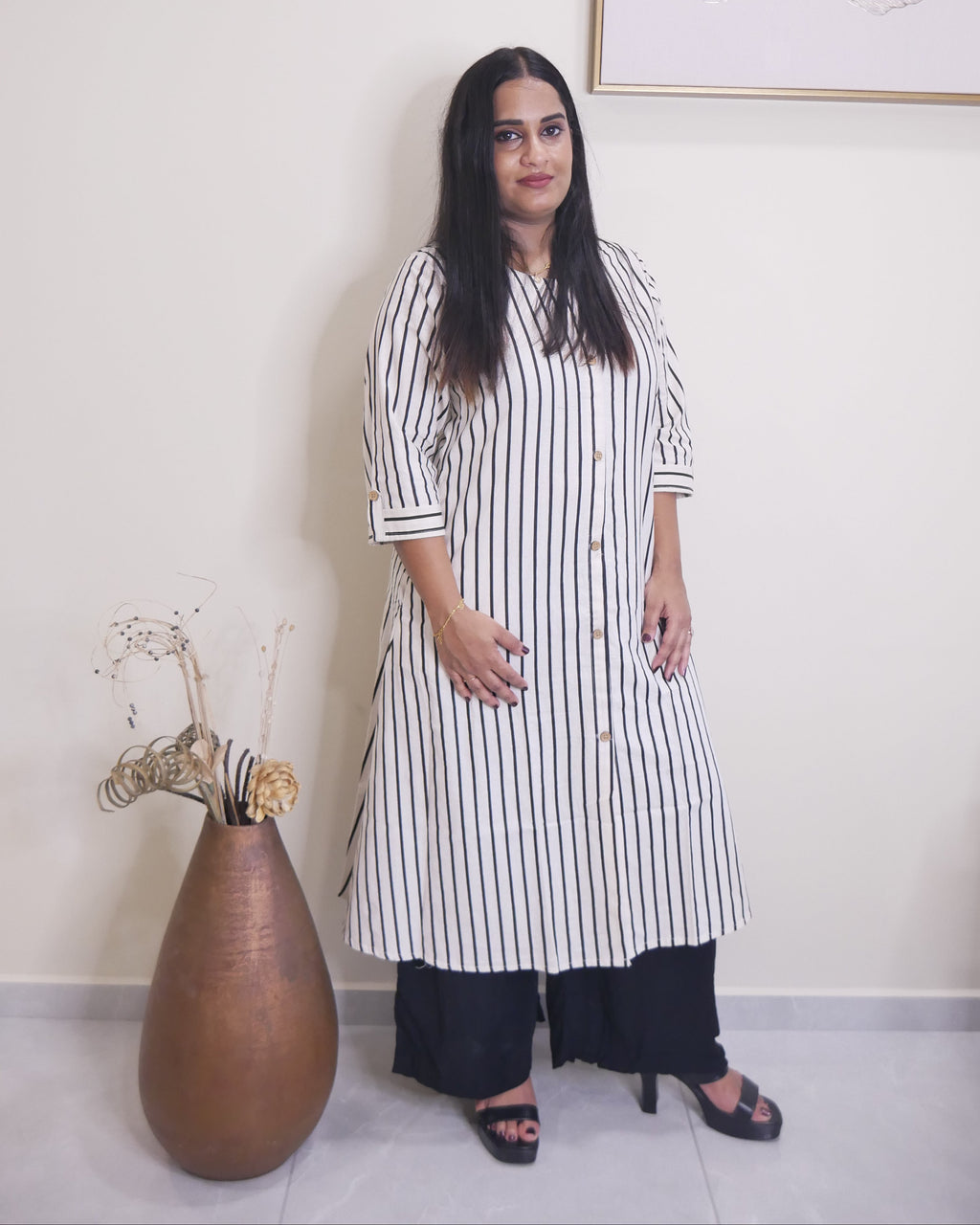 A woman stands indoors wearing a long black-and-white vertically striped tunic dress with elbow-length sleeves, paired with black wide-leg pants and black heeled sandals. She is posed in front of a plain off-white wall with a framed abstract print and a large brown vase with dried stems on the floor.