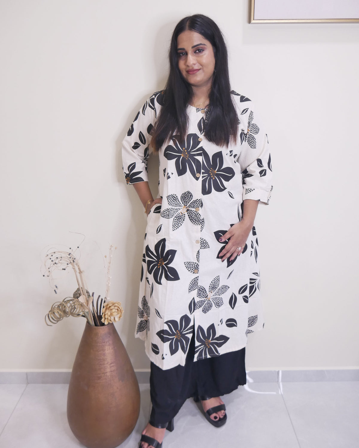 A woman stands indoors wearing a white long kurta with a black and gray floral print, paired with black pants and black sandals. She is posed next to a large brown ceramic vase with dried decorative stems against a plain off-white wall.