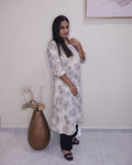A woman modeling a long-sleeved, knee-length kurta with a gray floral pattern on a white base, paired with black pants and black sandals, standing in an indoor setting.