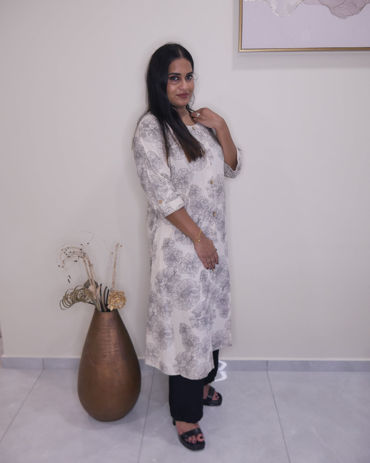 A woman modeling a long-sleeved, knee-length kurta with a gray floral pattern on a white base, paired with black pants and black sandals, standing in an indoor setting.