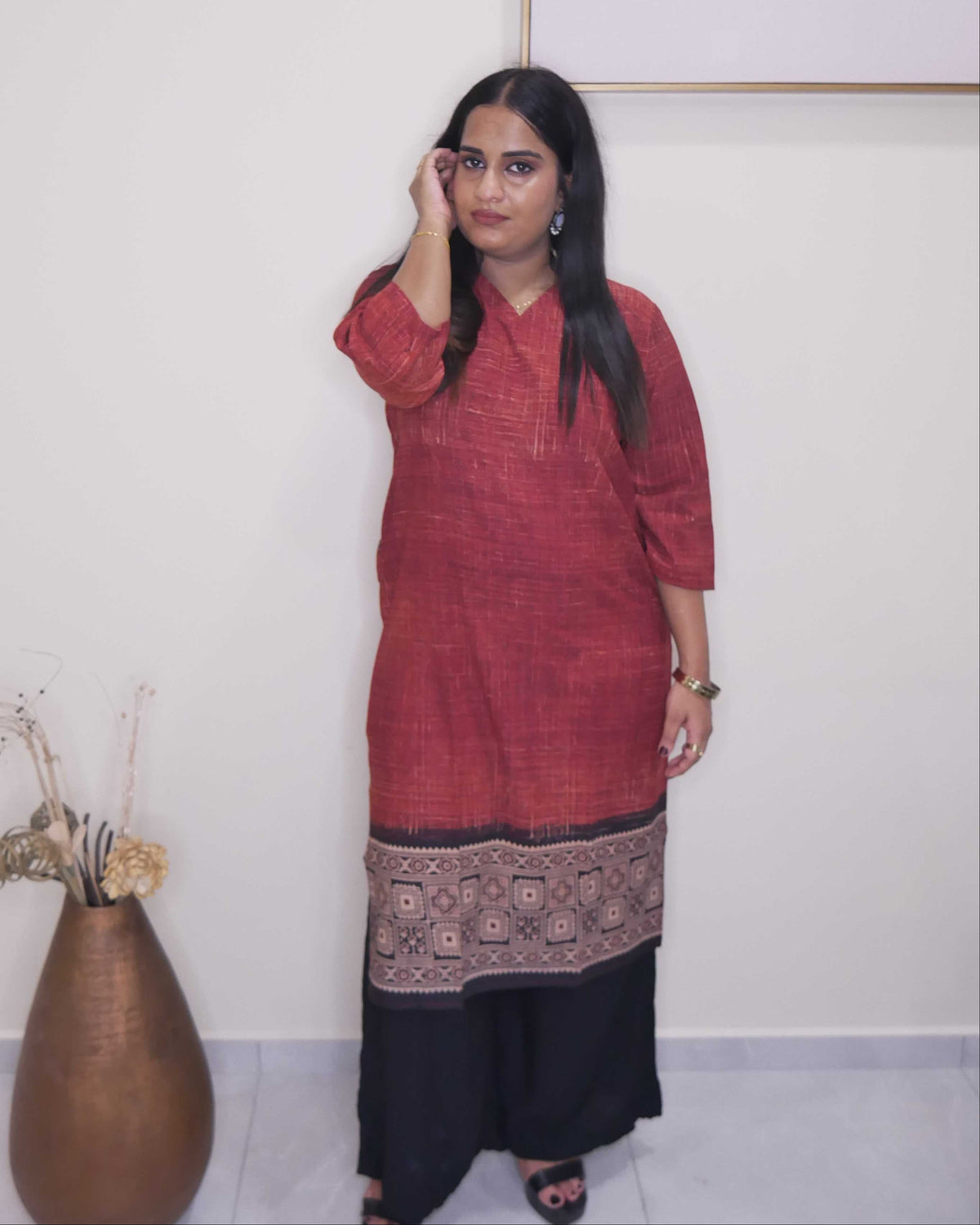 A woman stands indoors wearing a rust-red tunic with a dark patterned hem and black wide-leg pants, posed next to a large brown vase against a white wall.