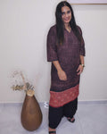 A woman modeling a dark plum purple textured kurta with a red patterned hem, paired with black pants, standing in an indoor setting next to a large brown vase.