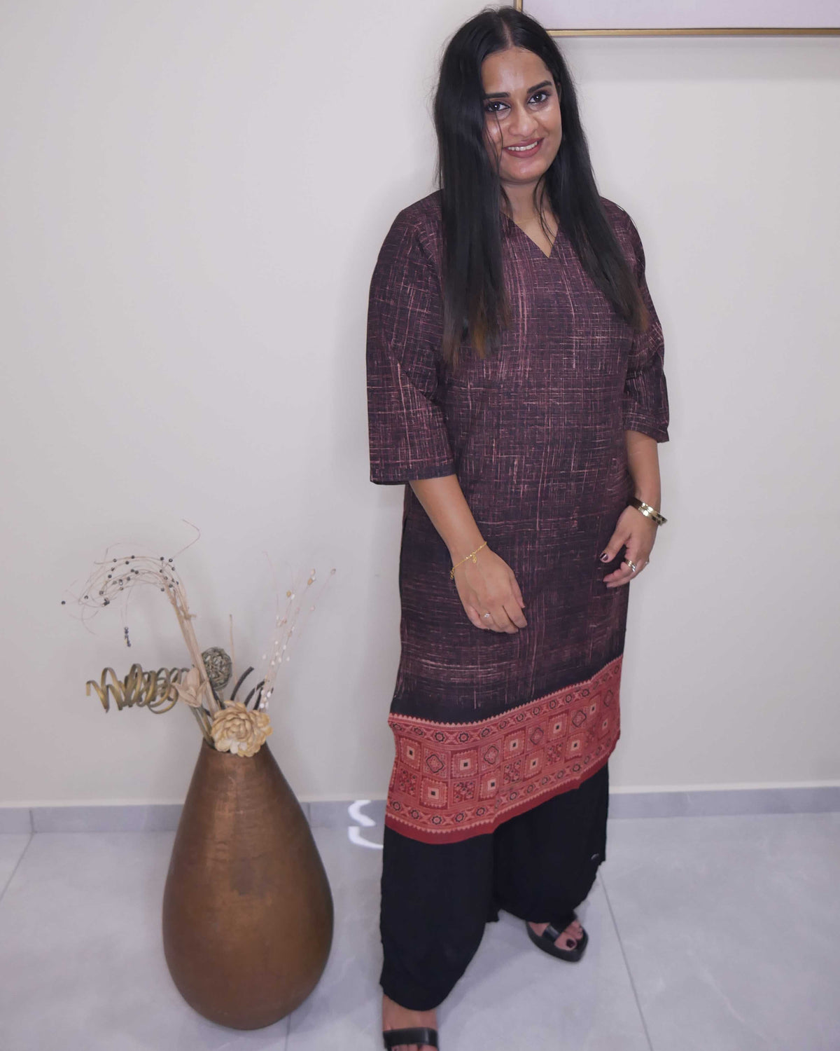 A woman modeling a dark plum purple textured kurta with a red patterned hem, paired with black pants, standing in an indoor setting next to a large brown vase.