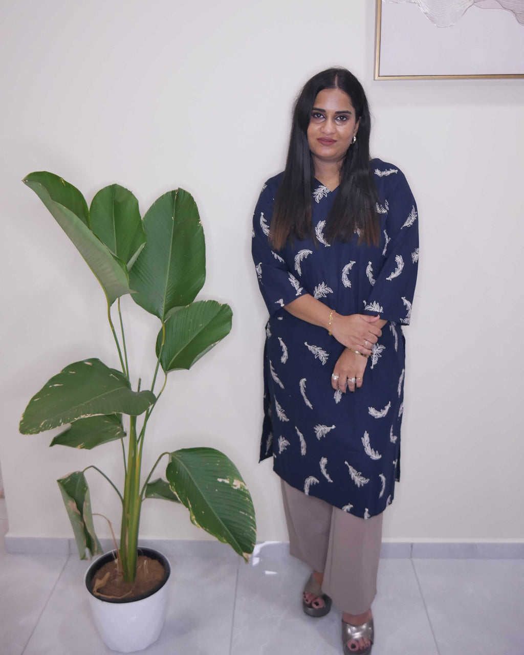 A woman stands indoors next to a large potted plant, wearing a navy blue tunic with a white abstract pattern and beige trousers.
