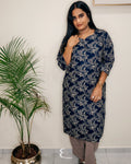 A woman modeling a navy blue and gray floral printed kurta with beige pants, standing in an indoor setting next to a potted palm plant against a plain wall.