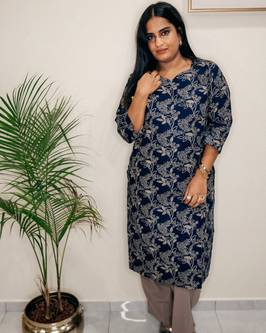 A woman modeling a navy blue and gray floral printed kurta with beige pants, standing in an indoor setting next to a potted palm plant against a plain wall.