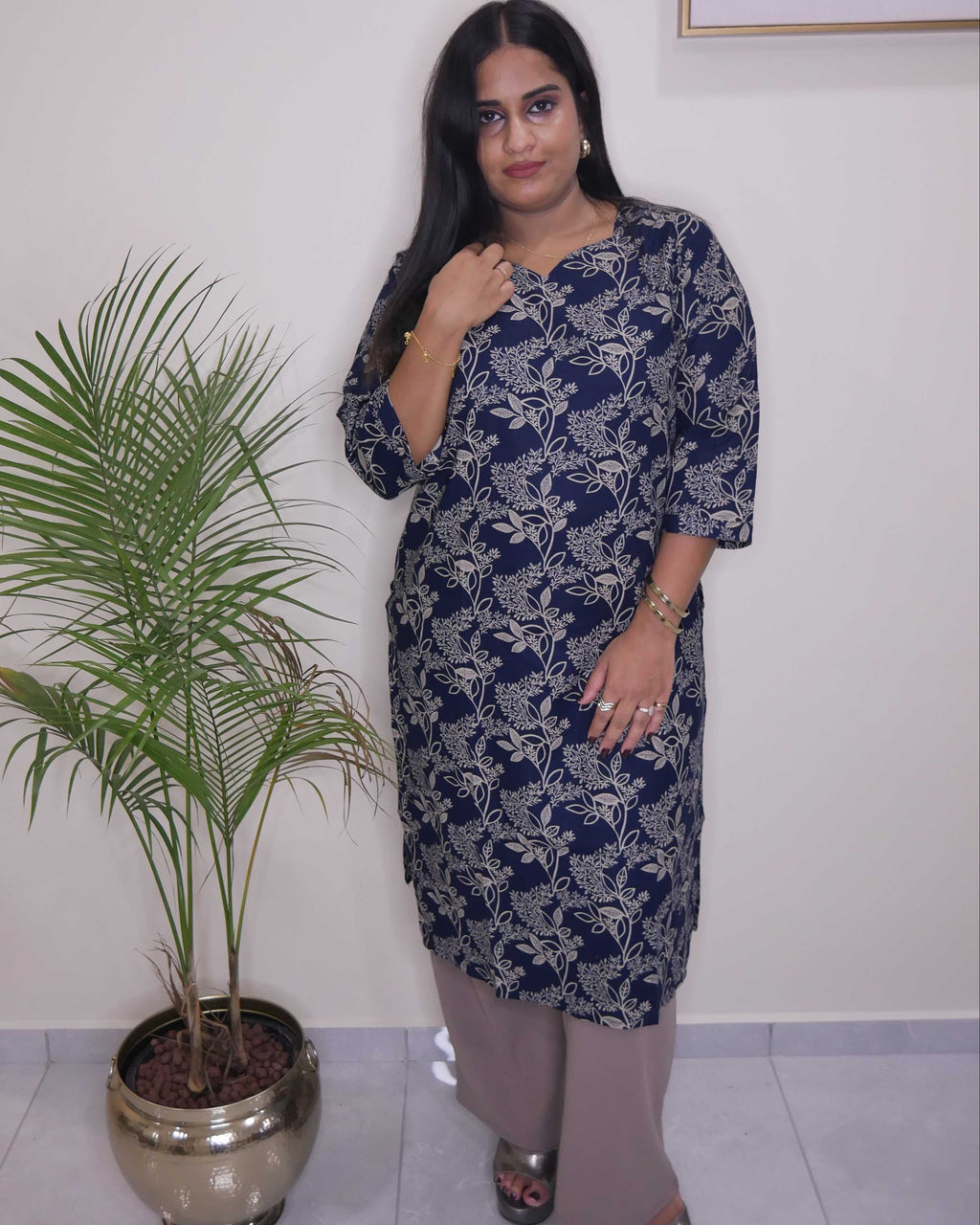 A woman modeling a navy blue kurta with a white floral pattern, paired with beige trousers and silver sandals, standing in an indoor setting next to a potted palm tree.