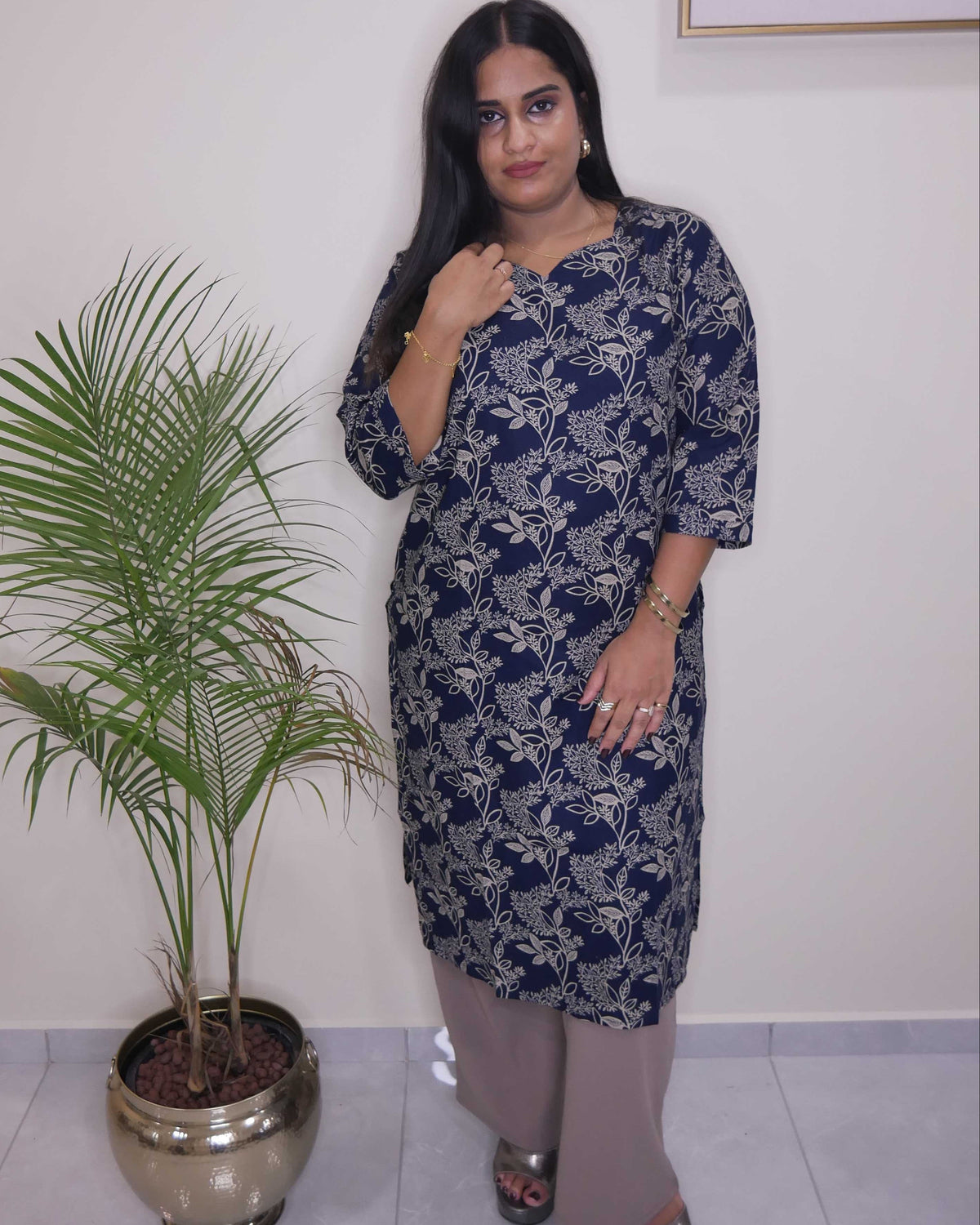 A woman modeling a navy blue kurta with a white floral pattern, paired with beige trousers and silver sandals, standing in an indoor setting next to a potted palm tree.