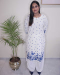 A woman modeling a white long kurta with blue floral and geometric patterns, paired with white wide-leg pants, standing in a room next to a potted palm tree.